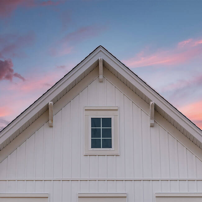 New gable with decorative brackets, white vertical vinyl lap siding with square attic window on a newly built home in America colorful sunset background