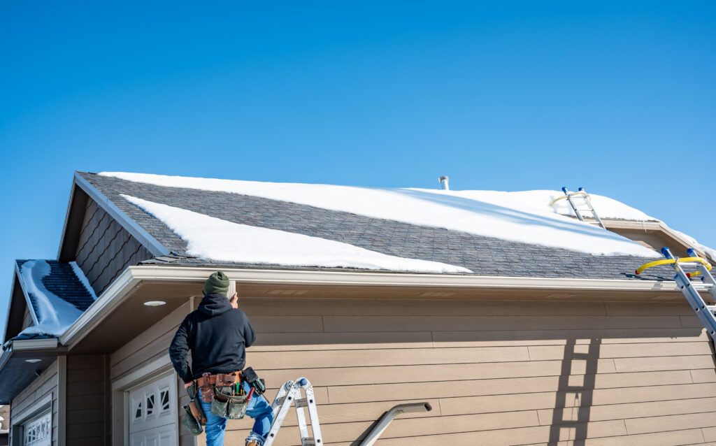 A contractor is installing gutters on a home. The shingled roof has patches of snow on it.