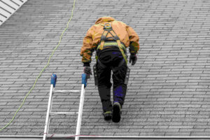 A man wearing a yellow jacket and safety gear is walking on a sloped gray shingled roof.