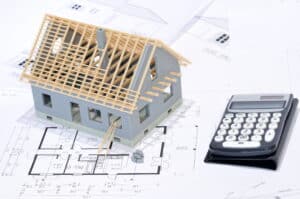 A model of a home with wood decking for a new roof sits on blueprints. A calculator sits next to the model home.